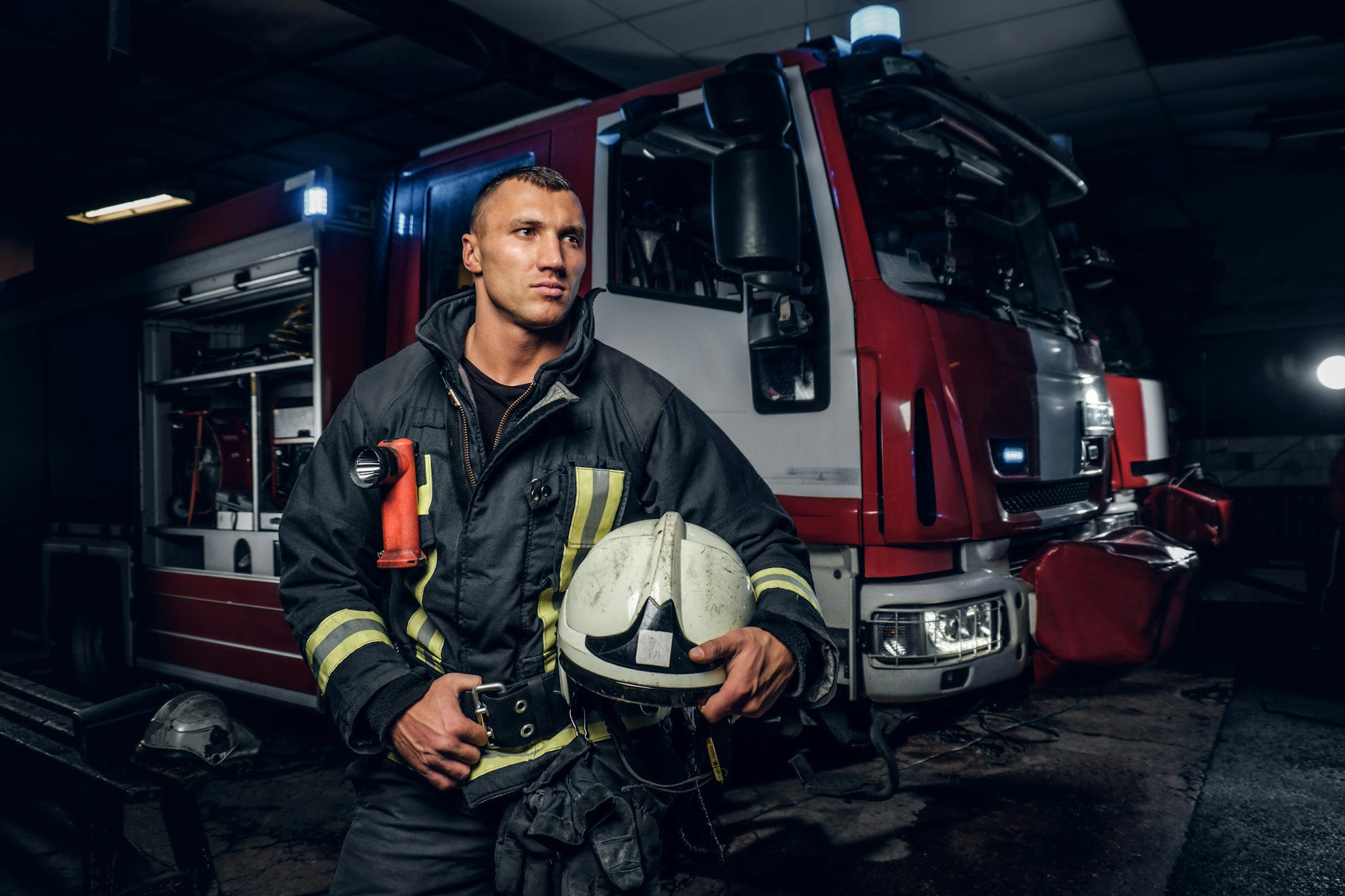 Firefighter in protective uniform standing next to a fire engine in a fire department garage, showcasing readiness and courage in emergency response.